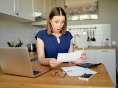 Woman in a kitchen looking shocked while reviewing a document, illustrating concerns about unexpectedly high water bills indicating potential plumbing leaks.