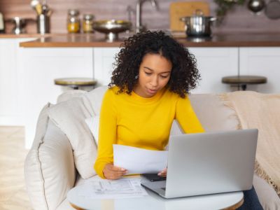 Woman in yellow sweater reviewing gas bill and documents on laptop at home, indicating potential gas leak concerns.